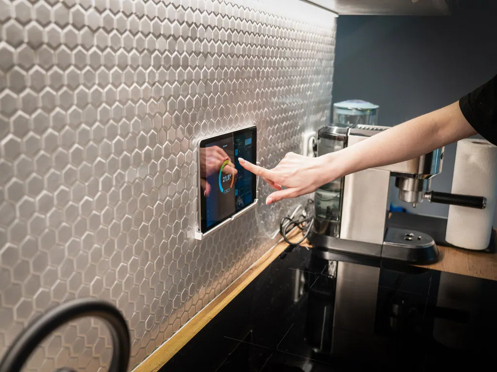 A hand tapping a wall-mounted touch panel on a tiled kitchen wall