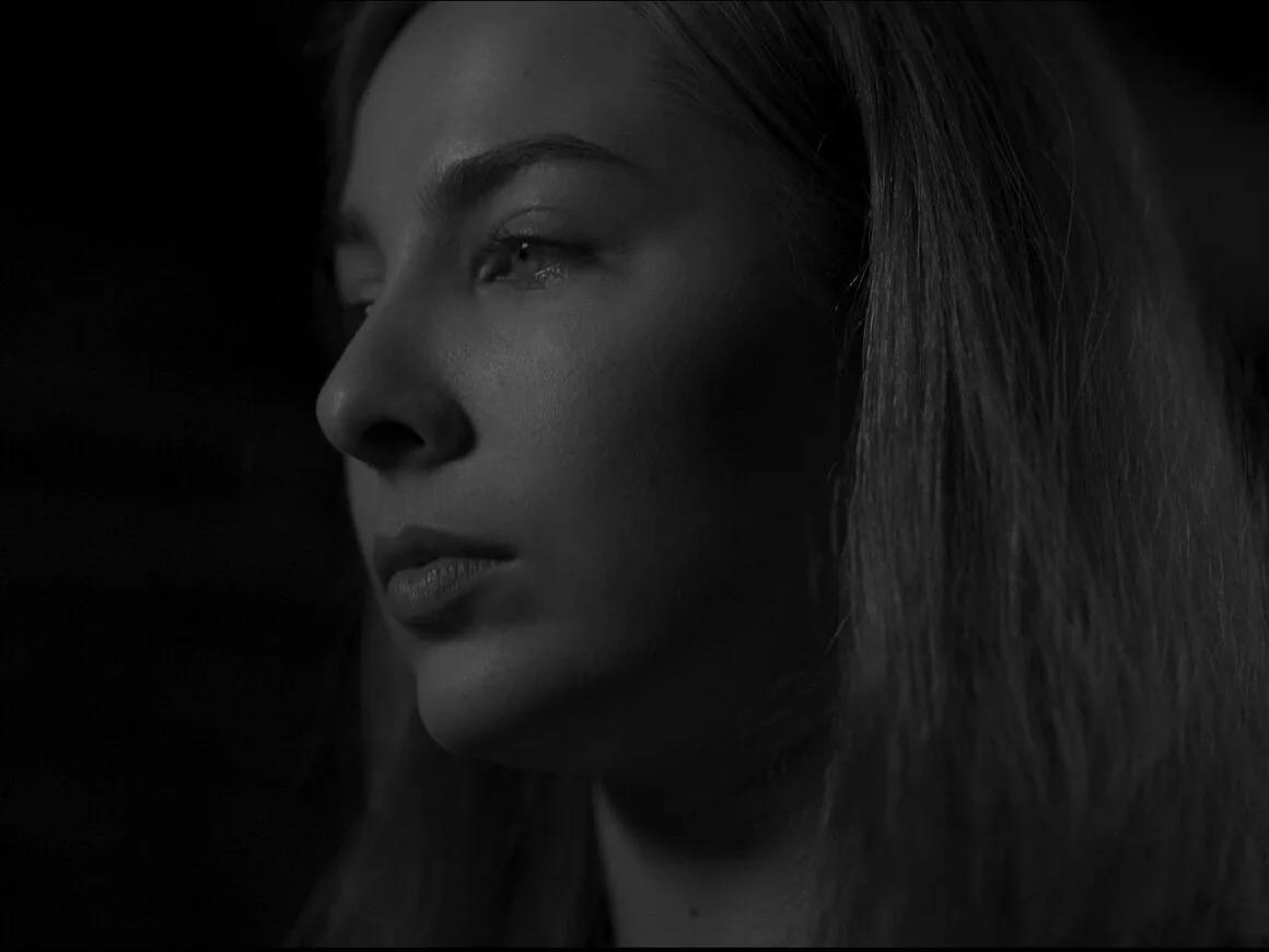Black-and-white film still: a young woman in profile, lit from one side against a dark room