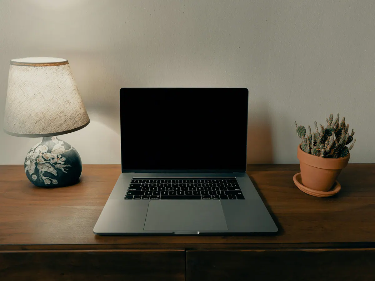 A laptop on a walnut desk between a linen table lamp and a small potted cactus