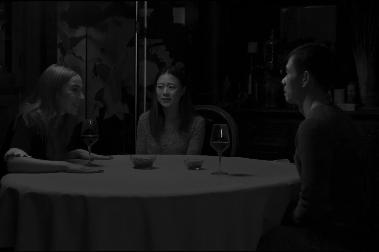 Film still from Exsanguination, in black and white: three people talk across a round dinner table set with wine glasses