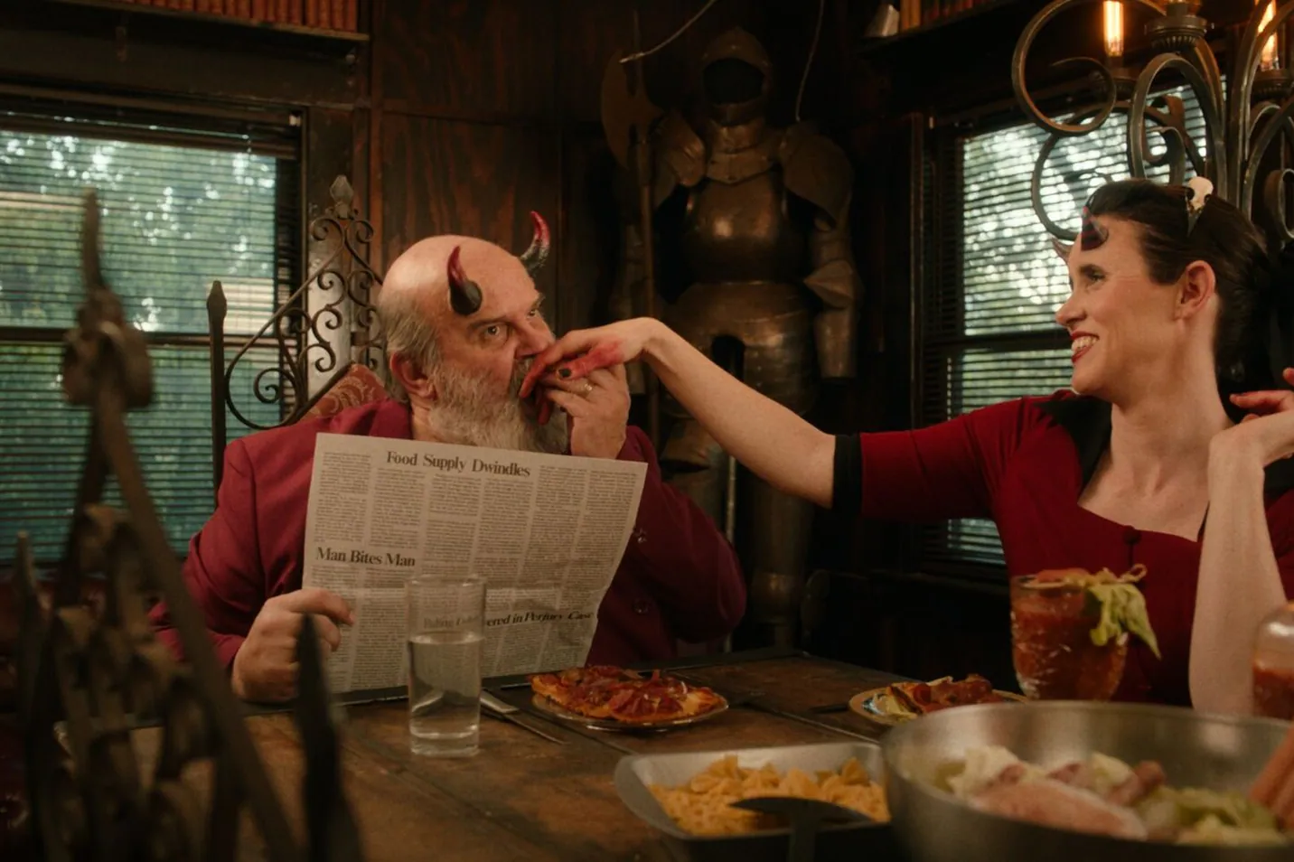 Film still from The Morningstars: a horned couple at a cluttered breakfast table, he behind a newspaper, she reaching across to him