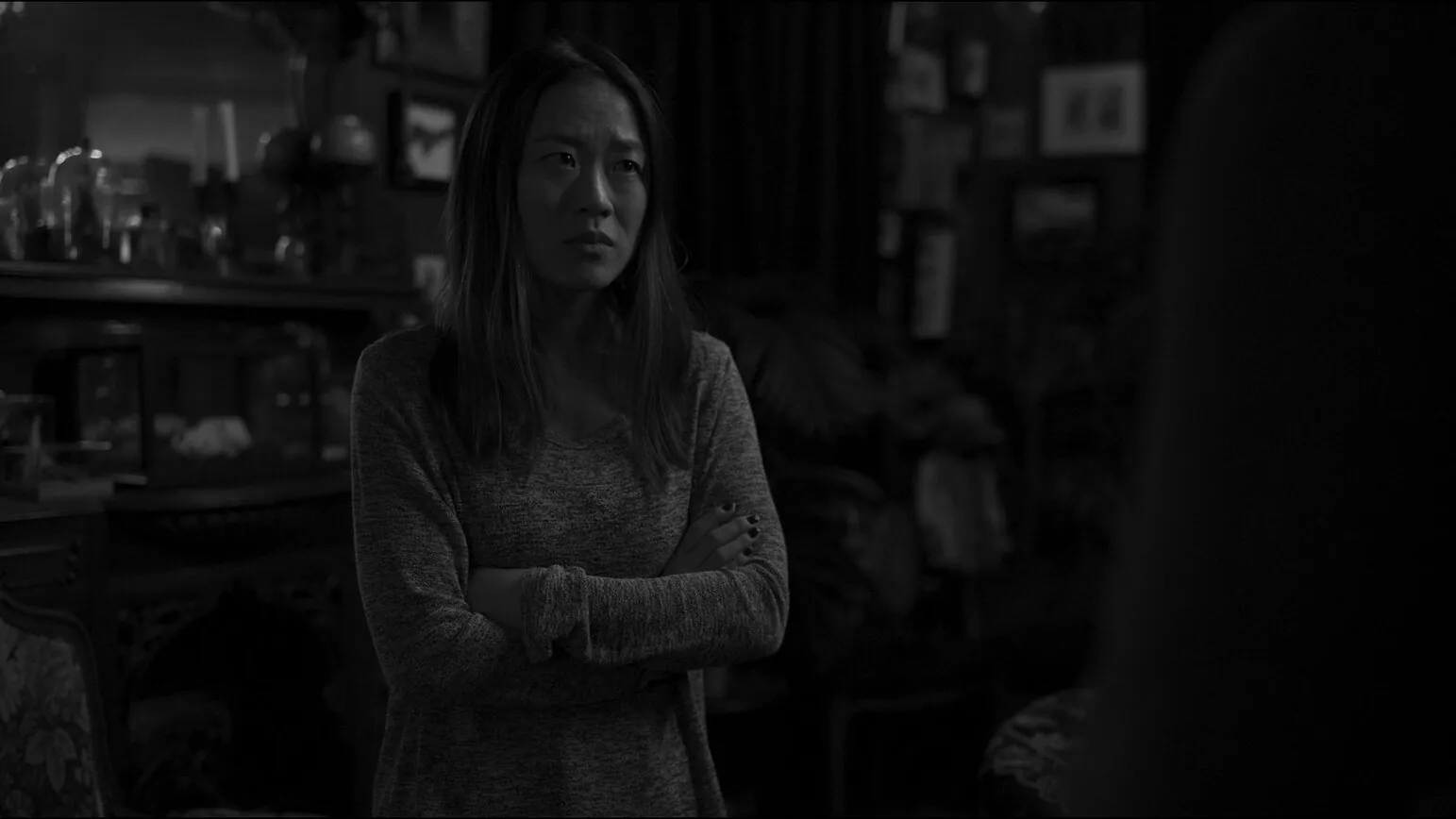 Film still from Exsanguination, in black and white: a woman stands with her arms crossed in a cluttered, dimly lit room