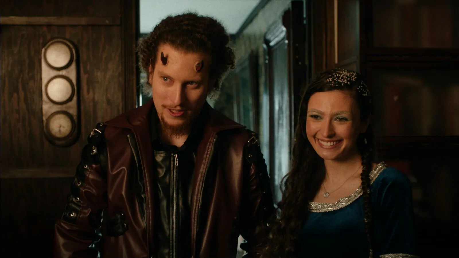Film still from The Morningstars: a horned young man in a leather coat grins beside a smiling young woman in a blue gown