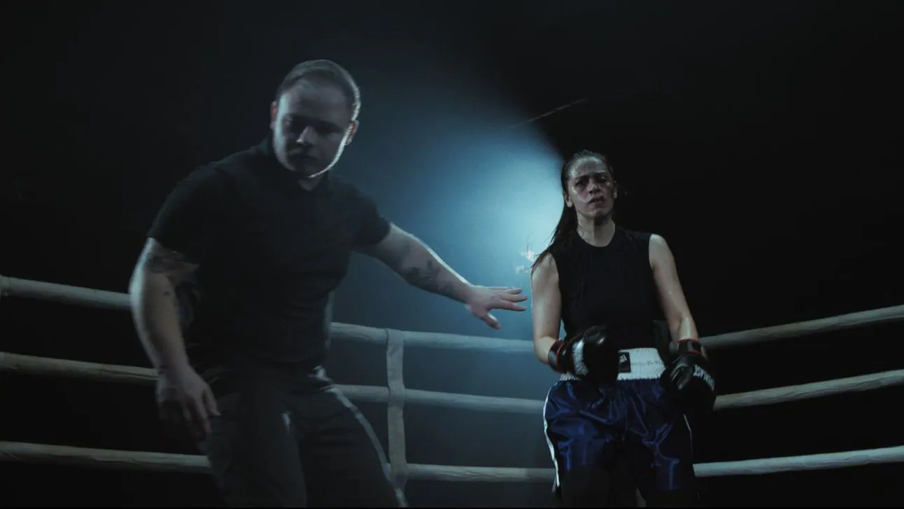 Film still from Piroba: a referee holds out an arm as a boxer in blue shorts stands against the ropes under a cold spotlight