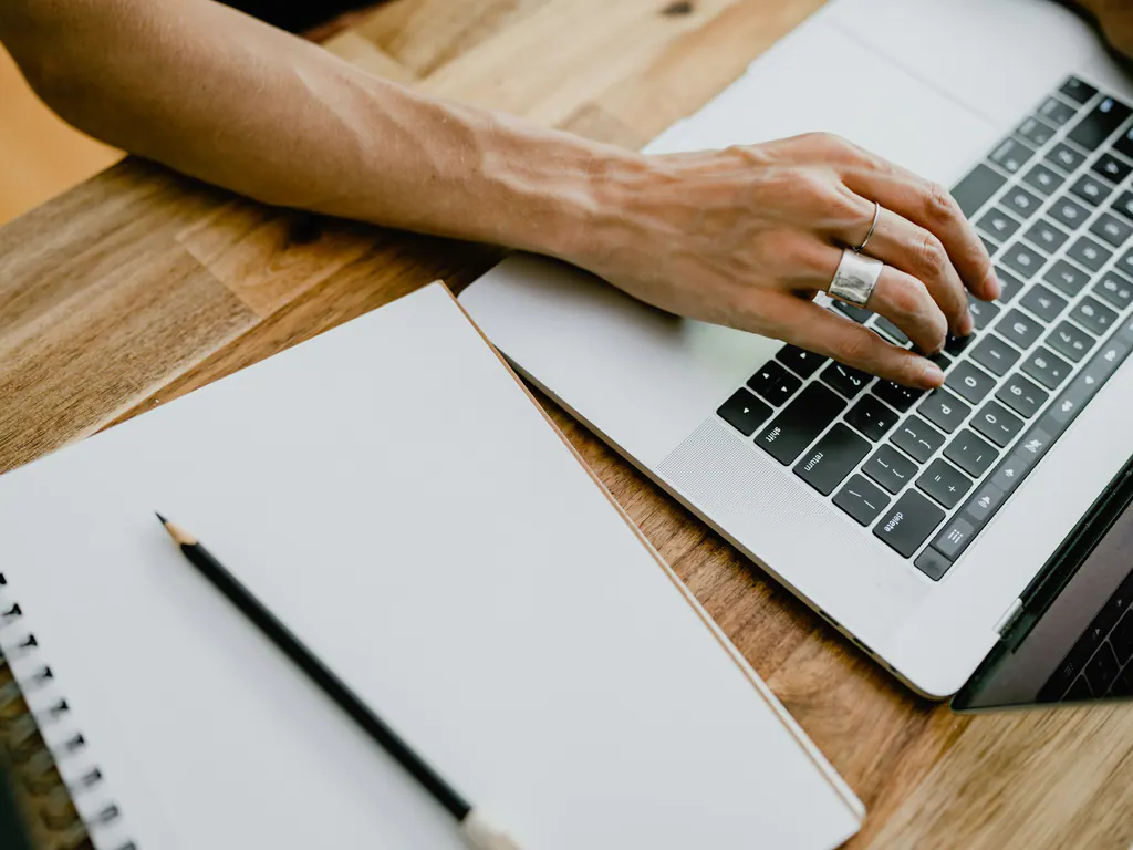 A hand on a laptop keyboard beside an open notebook and a pencil on a wooden table